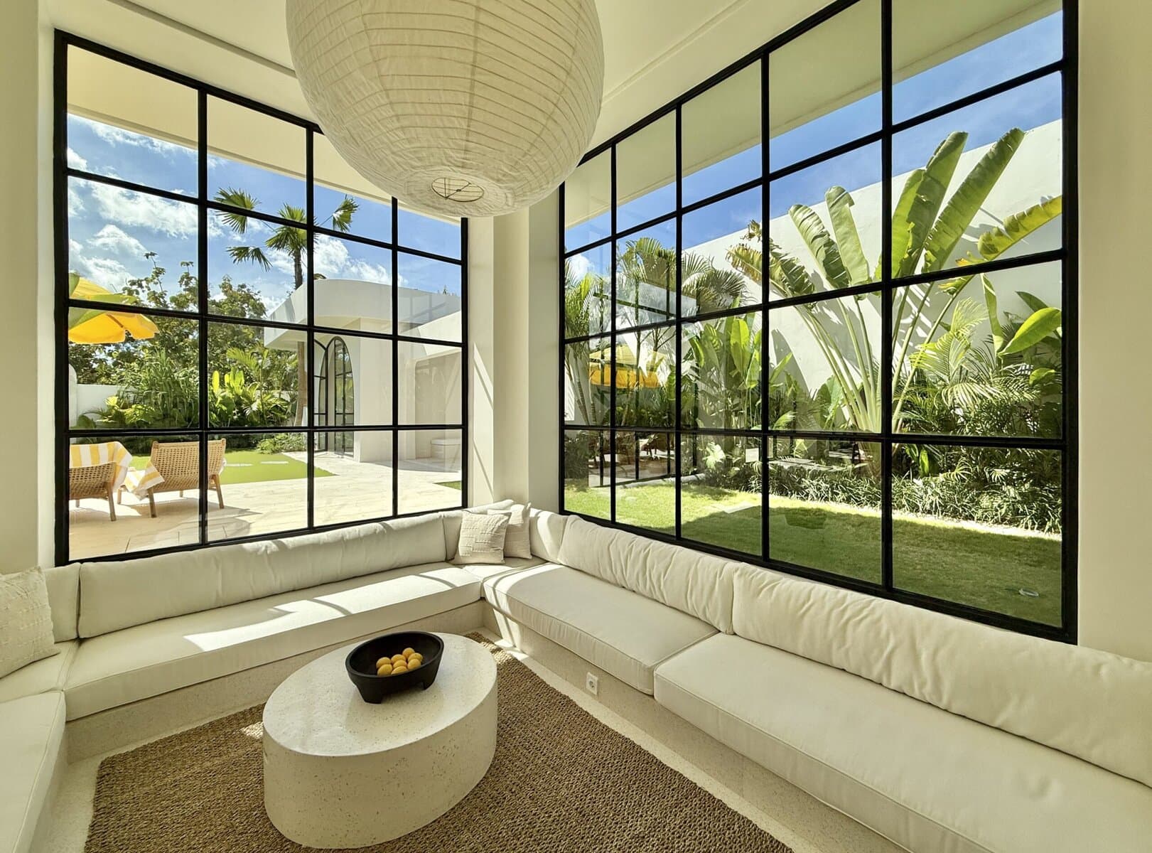Modern tropical interior with large glass panels, white sofa, vibrant outdoor greenery.
