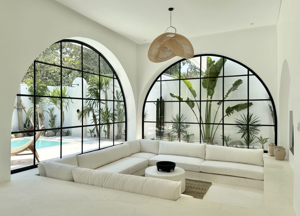 Bright living room featuring white seating, black-framed arched windows, and tropical garden views.