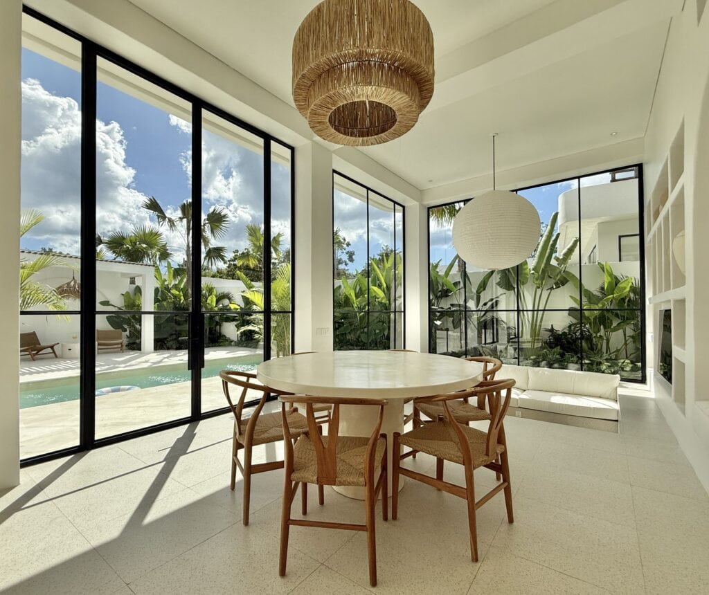 Modern tropical living space with panoramic glass walls, natural light, and lush greenery outside.