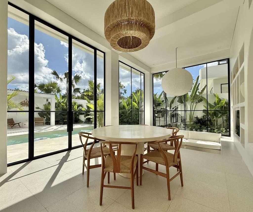 Modern tropical living space with panoramic glass walls, natural light, and lush greenery outside.