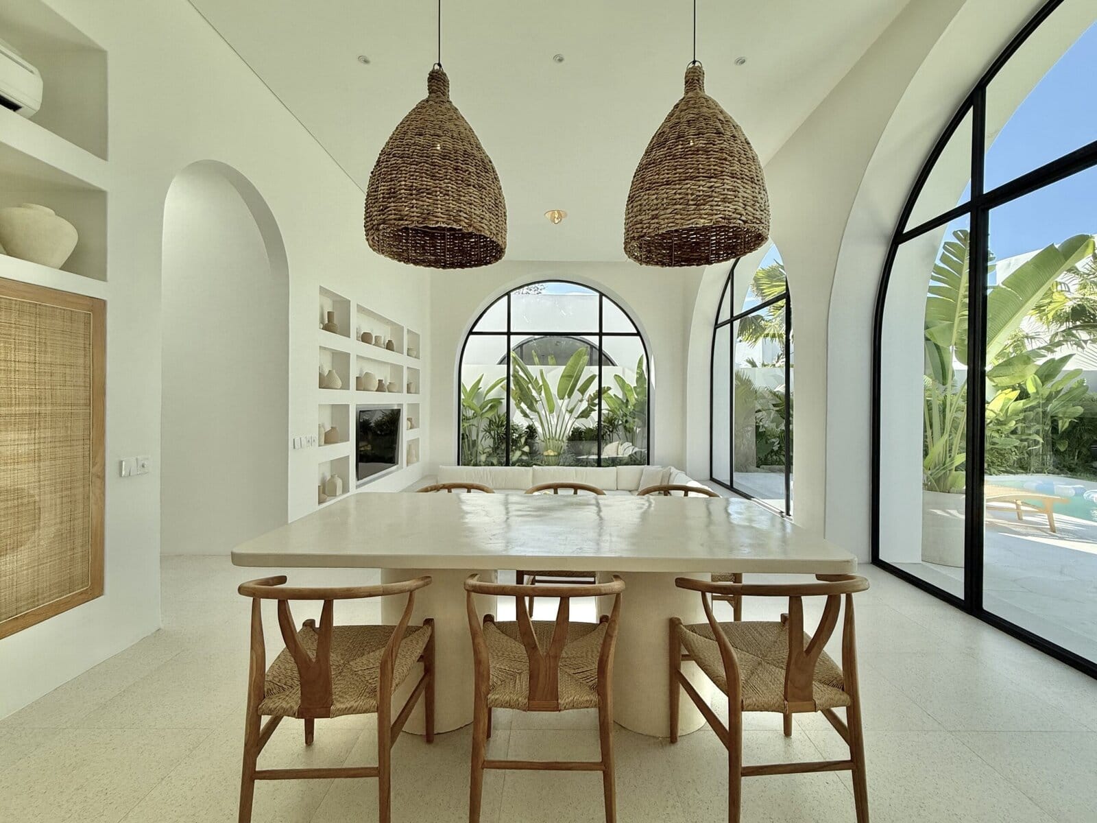 Elegant indoor dining area with curved glass windows, tropical lush greenery outside, woven pendant lights, and minimalist furniture.