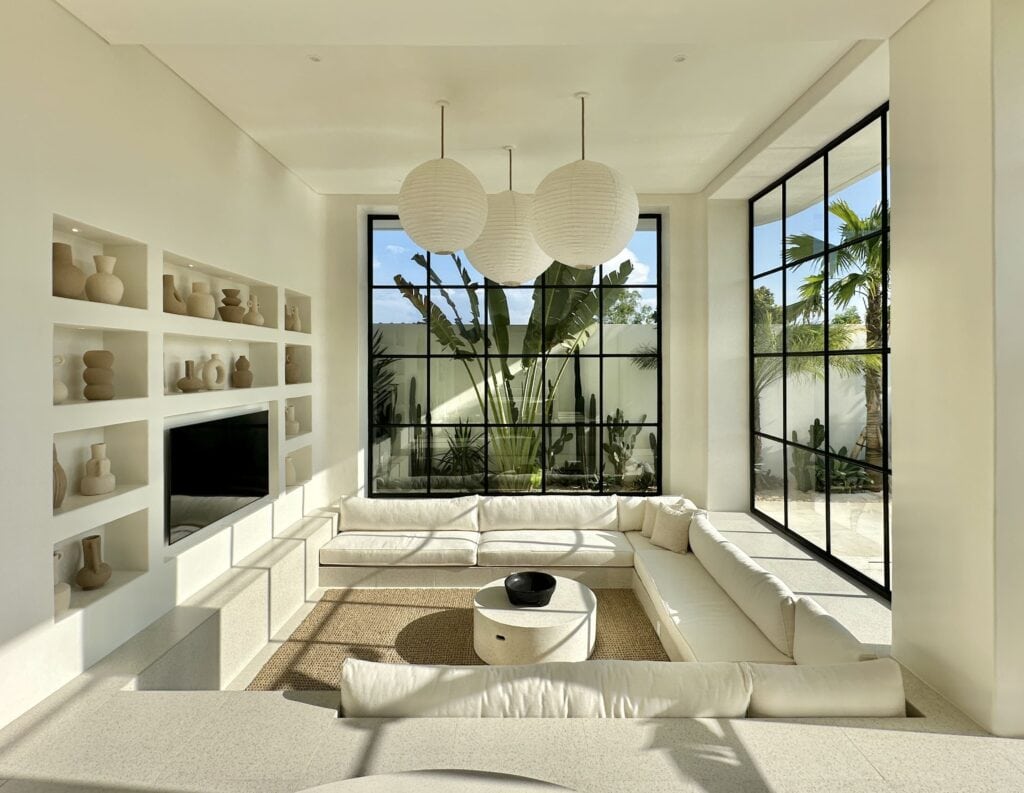Bright, spacious living room in a luxury Bali villa featuring minimalist decor, large black-framed windows, and tropical greenery outside.
