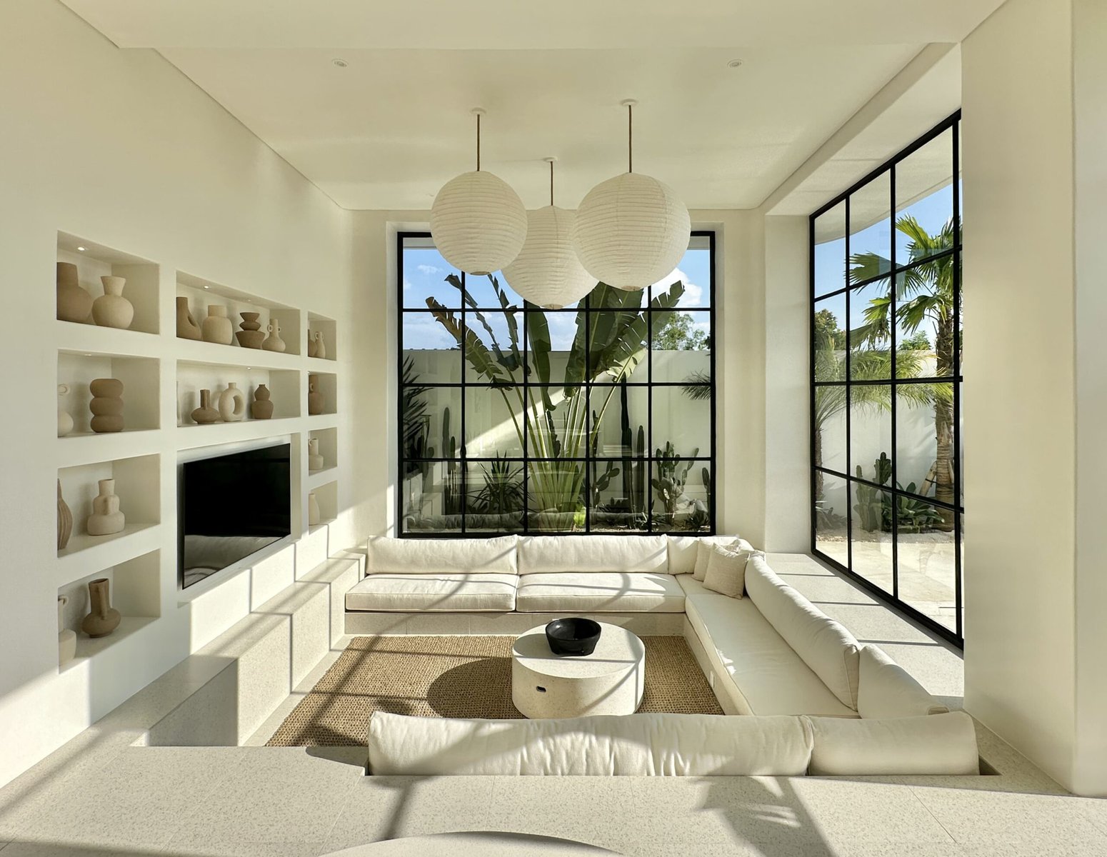Bright, spacious living room in a luxury Bali villa featuring minimalist decor, large black-framed windows, and tropical greenery outside.