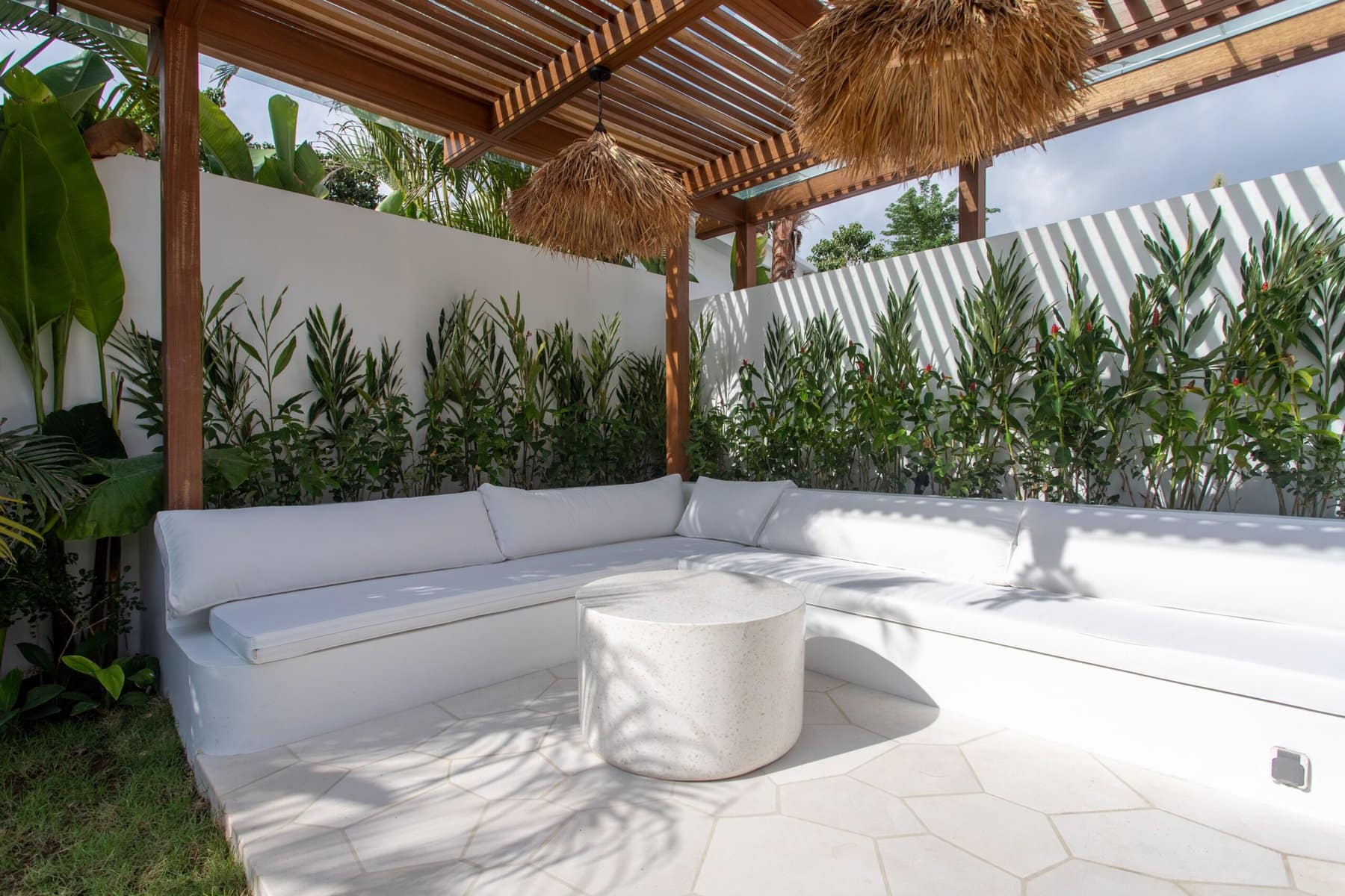 Elegant white outdoor sofa with tropical plants and shaded pergola outdoors.