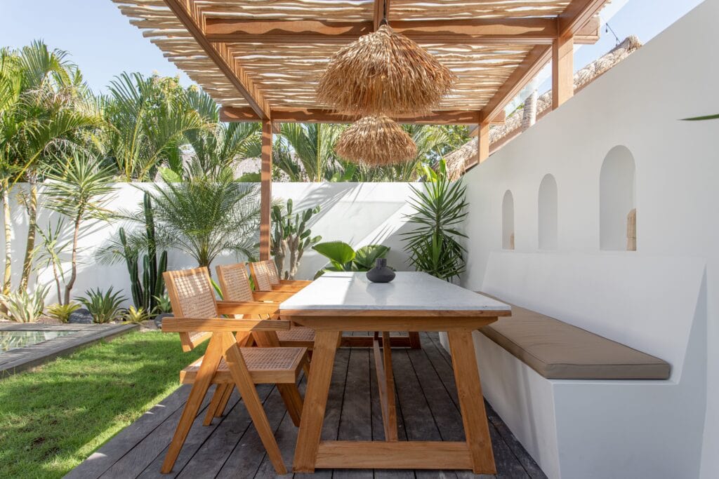 Bright outdoor dining space with wooden table, shaded by natural thatched lamps, surrounded by lush greenery.