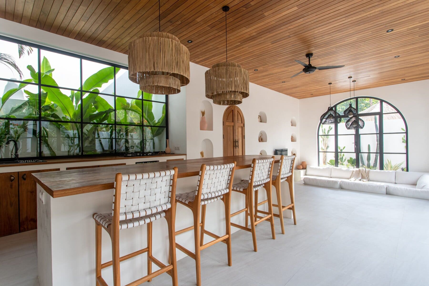 Bright, airy living space with modern minimalist design, wooden accents, and large tropical windows.