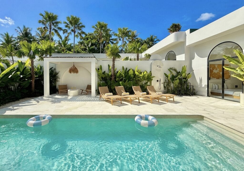 Modern tropical villa with a turquoise swimming pool, wooden deck featuring rattan lounge chairs, and a lush garden of palm trees under a bright, cloud-dotted sky at noon.