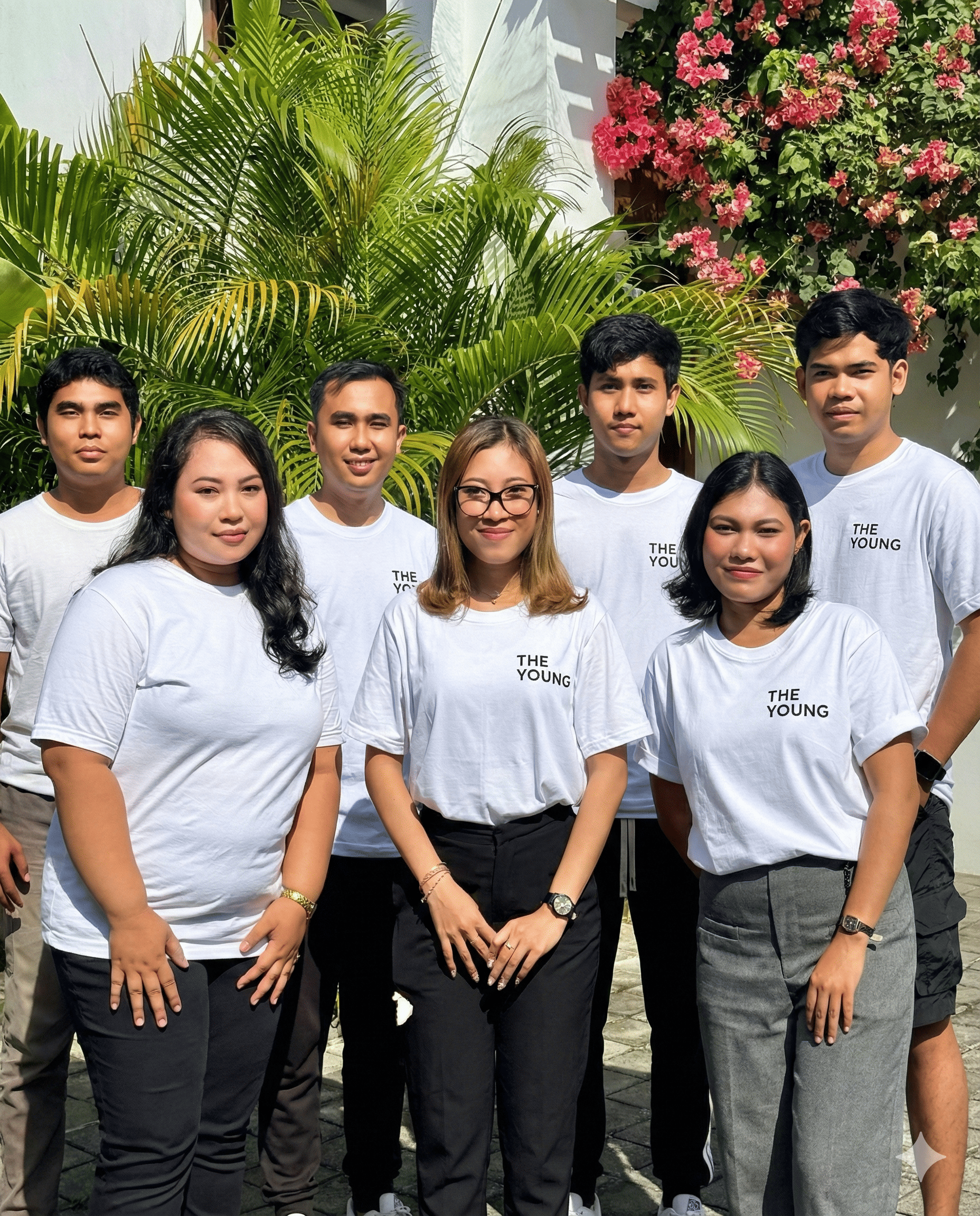Group of seven young adults standing outdoors wearing matching white t-shirts with 'THE YOUNG' logo on the chest, posed in front of tropical greenery and pink flowers with soft natural lighting.